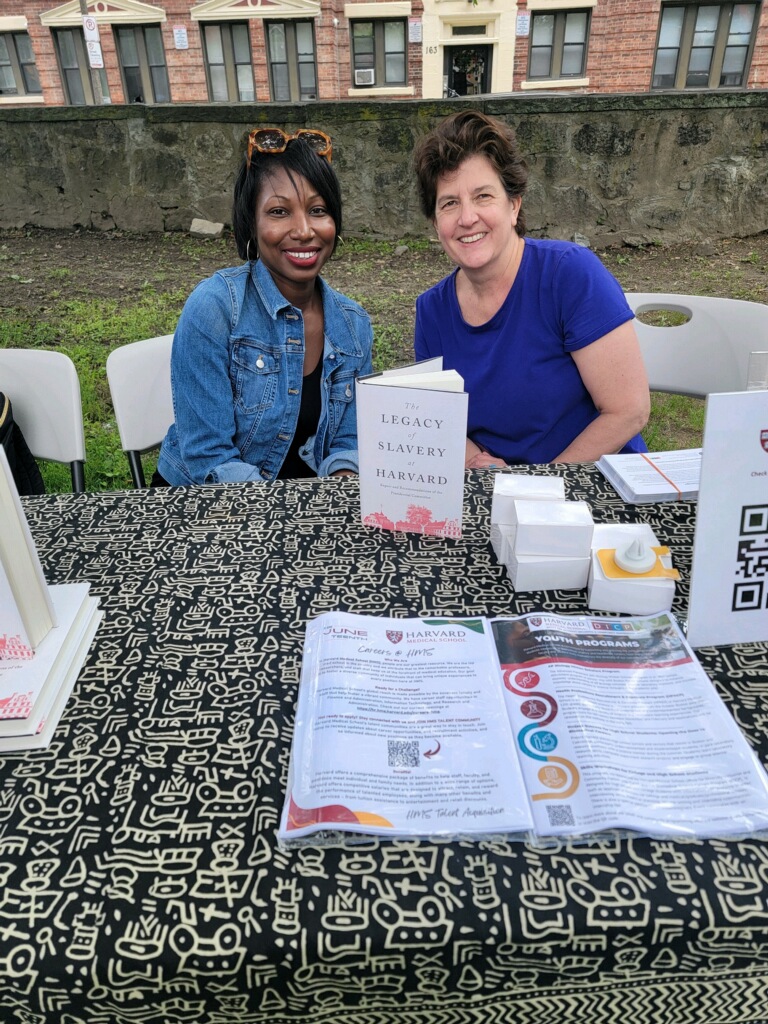 Michelle Keenan of DICP and Roeshana Moore-Evans from Legacy of Slavery at Harvard smile at the camera as they sit by a table with flyers promoting Harvard happenings.